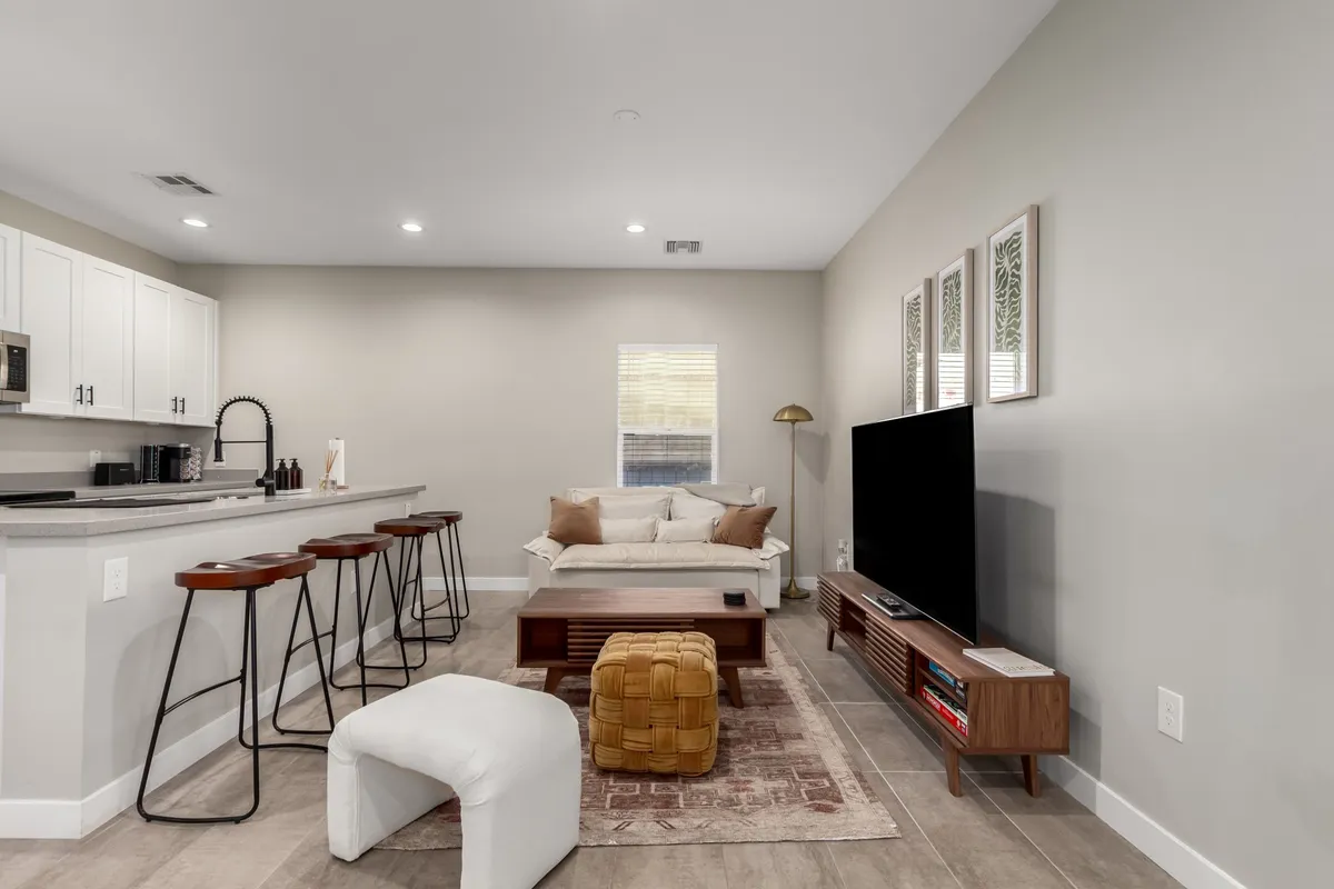This image provides a clear view of the open living area, featuring a comfortable sofa, a kitchen island with bar stools, a television stand with a TV, and warm decorative wall accents. The scene exudes a casual, inviting atmosphere ideal for group relaxation, movie nights, and easy conversation. The open layout hints at the living room's versatility and the connected spaces.
